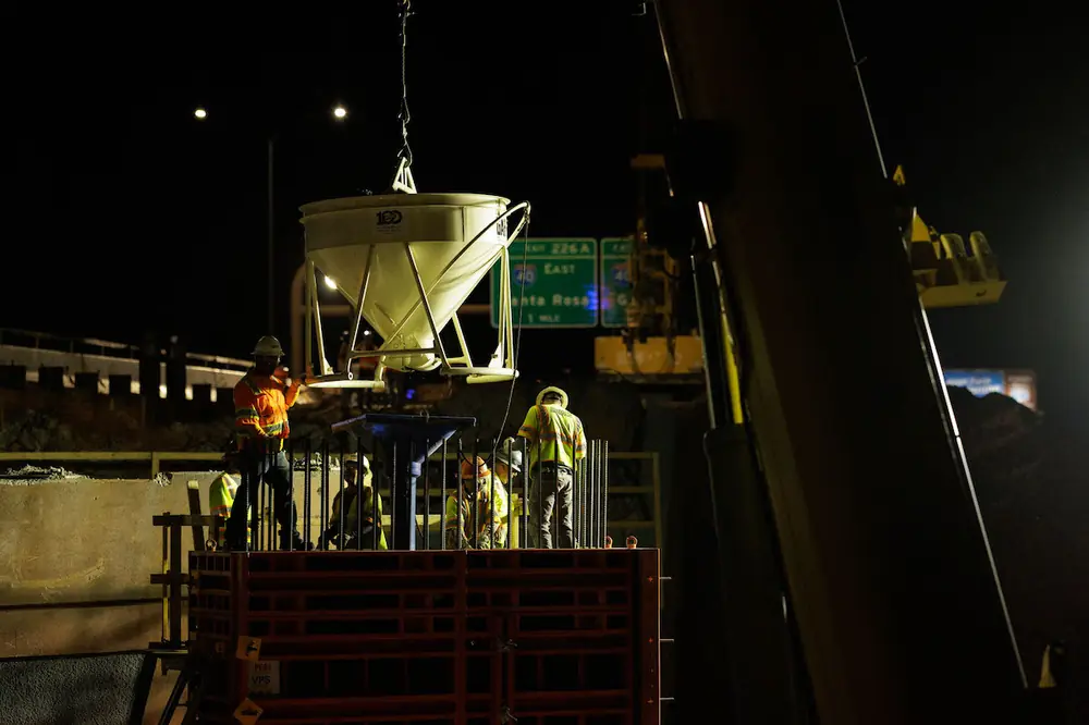 Crews stand on top of a bridge pier form along Comanche Road on the west side of I-25 as a bucket brings a load of concrete to the form.