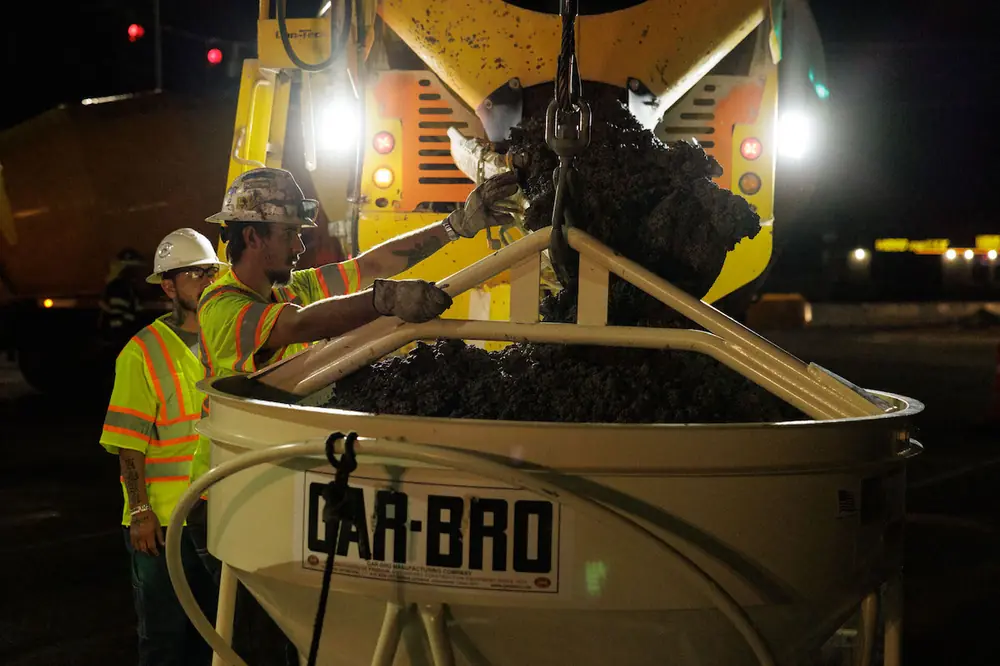 Two crewmembers fill a bucket, preparing the next load of concrete to be hoisted to the top of a bridge pier form on Comanche Road on the west side of I-25.