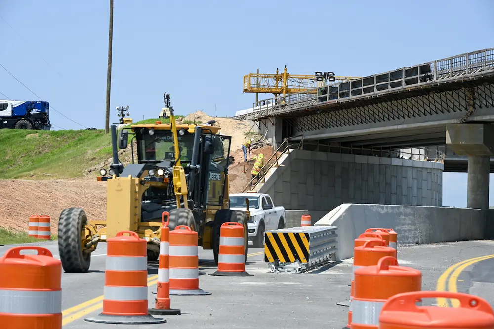 Contractors move equipment around the I-77 Exit 26 project site. In the background, the Exit 26 southbound on-ramp overpass is seen under construction.