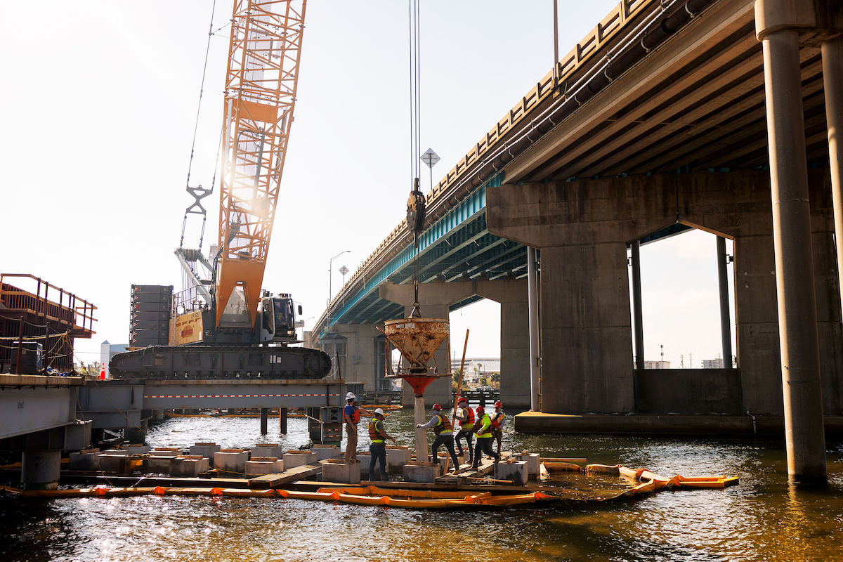 A crane positions materials during active bridge construction over the water, showcasing the complex marine logistics required for the Brooks Bridge Replacement.