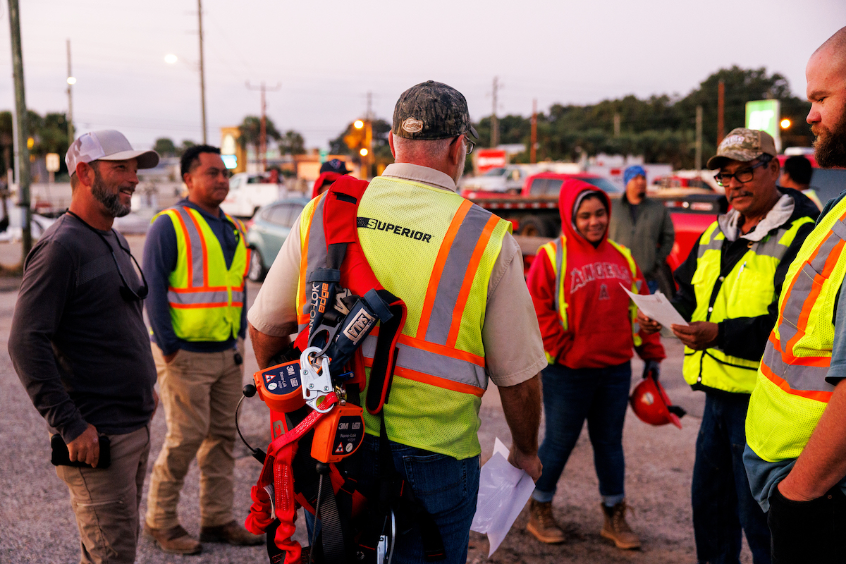 Superior Construction workers gather for an evening safety briefing during the Brooks Bridge Replacement Project, with a supervisor demonstrating proper equipment usage protocols.